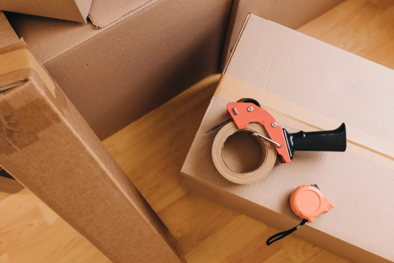 Umzugskartons mit Paketbandabroller und Maßband auf Holzboden.