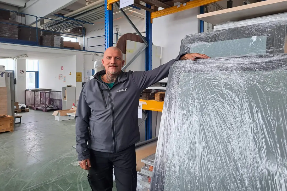 Ein lächelnder Reoffice-Mitarbeiter im Lager neben verpackten Büromöbeln.